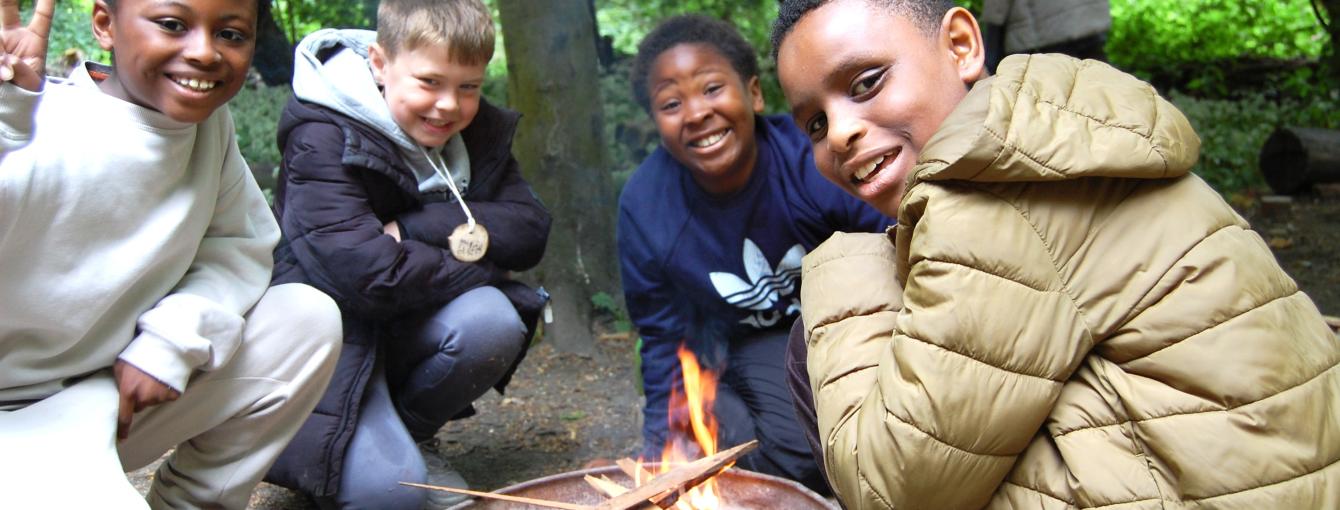 Three children in the woods, sitting round a campfire, smiling at the camera.
