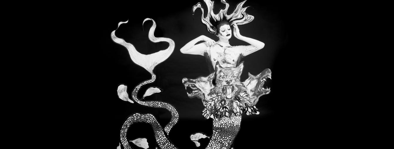 A monochromatic mermaid stands against a plain black backdrop. Her eyes are closed, hands framing her face and has long flowing hair. Her tail is long and spindly with three snarling dog heads placed at her waist.